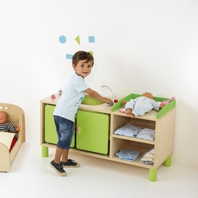 Móvel infantil de madeira clara com portas verdes e torneira de brinquedo, roupas e bonecos, e cama pequena para boneco ao lado.