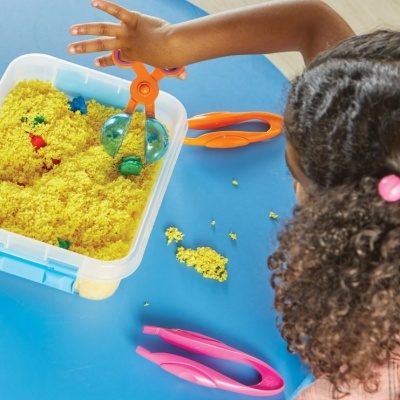 Recipiente com areia amarela granulada e pinças coloridas sobre mesa azul