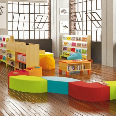 Espaço interior colorido com bancos, estantes de livros e janelas grandes em ferro.