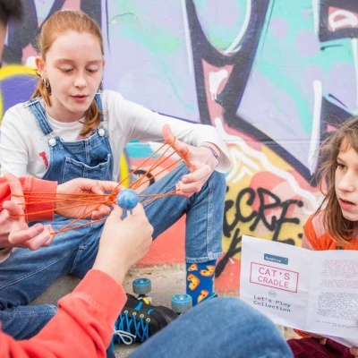 Três crianças a jogar com fio de malabarismo junto a uma parede com graffiti colorido.