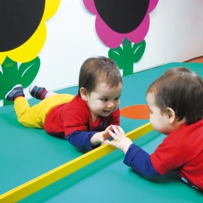 Bebé a olhar para o seu reflexo num espelho junto a uma parede decorada com flores coloridas