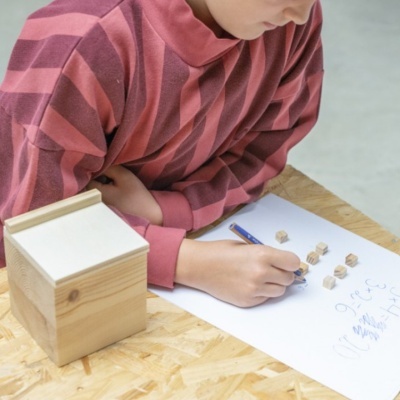 Pessoa com camisola riscada a escrever com lápis em papel sobre mesa de madeira, caixa de madeira ao lado
