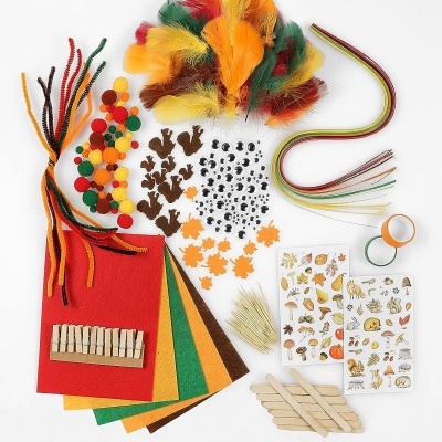 Materiais de artesanato coloridos organizados sobre superfície branca, incluindo feltro, plumas, contas, recortes de tecido e papel, palitos e fitas.