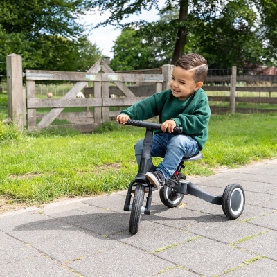 Criança num triciclo cinza escuro com guiador preto num passeio junto a relvado e cerca de madeira