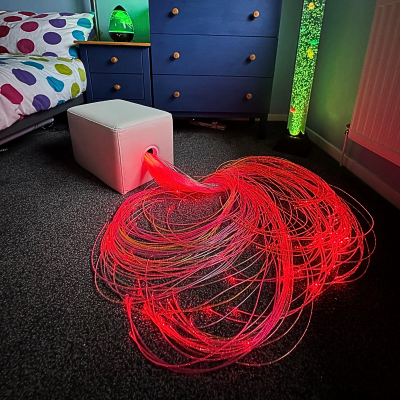 Cubo branco com fibras óticas vermelhas a sair, cama com colcha colorida e móveis azuis numa divisão com luz verde