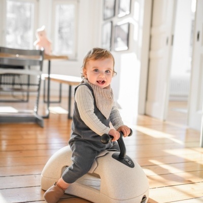 Criança sentada num brinquedo de montar bege com volante preto em madeira clara