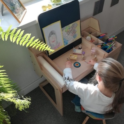 Criança sentada numa secretária de madeira com materiais de desenho e planta verde junto à janela