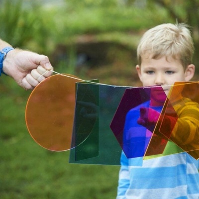Painéis coloridos em plástico translúcido com formas geométricas sendo segurados à frente de criança ao ar livre