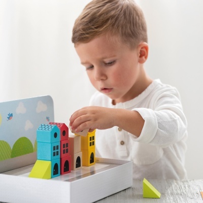 Criança a brincar com blocos de casas de madeira coloridas sobre uma superfície branca com fundo decorativo.