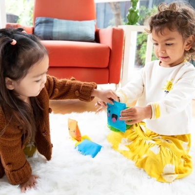 Duas crianças a brincar com sacos coloridos sentadas num tapete branco felpudo em sala com poltrona laranja.