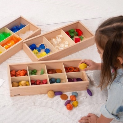 Criança a brincar com peças coloridas em caixas de madeira com divisórias sobre tapete branco.