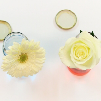 duas flores brancas em frascos de vidro com tampas douradas retiradas sobre superfície branca