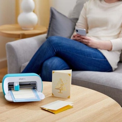 Máquina de corte Silhouette Cameo azul e branco sobre mesa com papéis e cartão amarelo, pessoa sentada no sofá ao fundo
