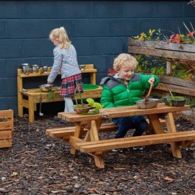 Mesa de madeira para piquenique infantil com bancos e crianças a brincar com plantas.