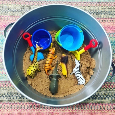 Balde metálico com areia e brinquedos de animais com pás coloridas, sobre tapete colorido