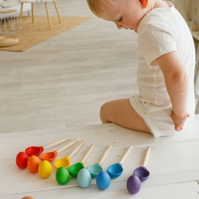 Brinquedos coloridos de madeira em forma de colheres sobre mesa branca, com criança ao lado.