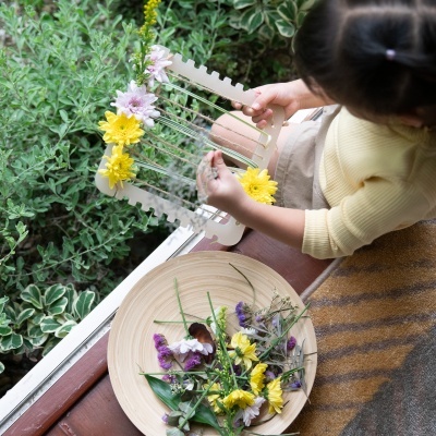 Criança segura moldura de madeira com flores coloridas ao lado de janela com plantas verdes