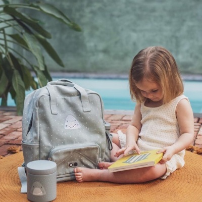 Mochila cinza claro com padrão de pontos e lancheira térmica cinza ao lado de criança loira sentada no chão lendo livro amarelo.