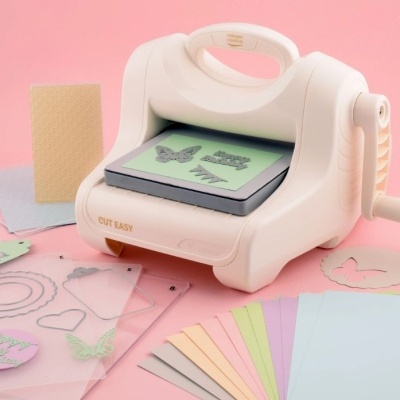 Máquina de corte manual branca CUT EASY com papéis coloridos e moldes transparentes sobre superfície rosa