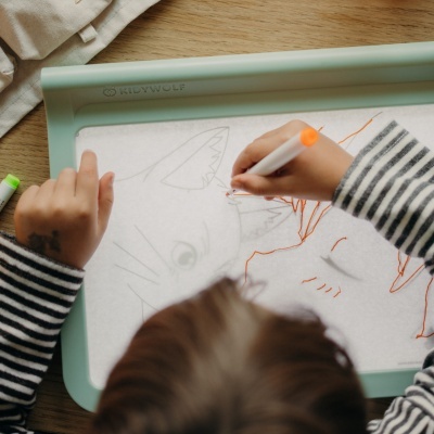 Mesa de desenho magnética verde com superfície branca e desenho de gato sendo feito por crianças com caneta laranja