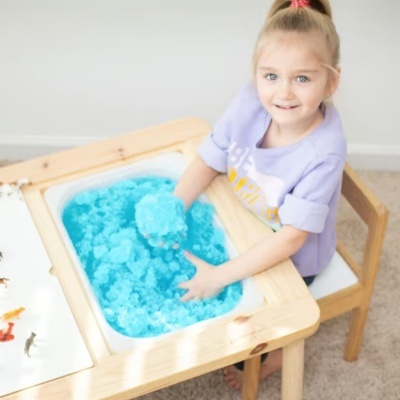 Criança sentada numa mesa de madeira com recipiente de areia azul e figuras de dinossauros