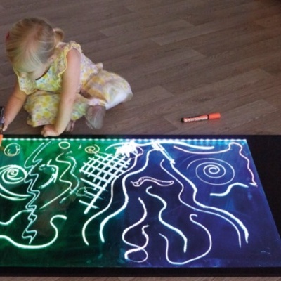 Criança a desenhar numa mesa digital luminosa sobre pavimento de madeira clara