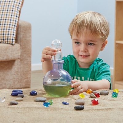 Criança segurando frasco transparente com líquido sobre tapete com pedras coloridas e lisas.