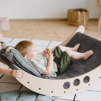 Criança deitada em cama curva de madeira com almofada preta às riscas, com outra criança a empurrar, em sala com tapete quadriculado
