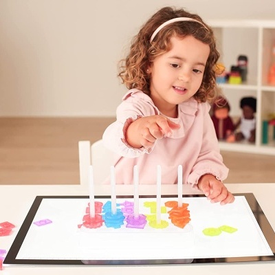 Criança a brincar com brinquedo colorido de plástico numa mesa branca, com estante e bonecos ao fundo