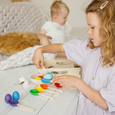 Criança a brincar com colheres e bolas coloridas numa mesa branca.