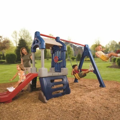 Parque infantil com escorrega, baloiços e painel giratório, crianças a brincar e mulher a observar no exterior