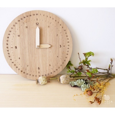 Relógio redondo de madeira clara com ponteiros às 12 horas, decorado com raminhos e flores secas numa mesa clara.