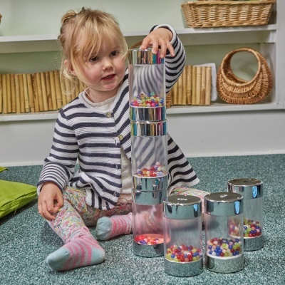 Criança a empilhar copos transparentes com bolinhas coloridas num chão verde com móveis ao fundo