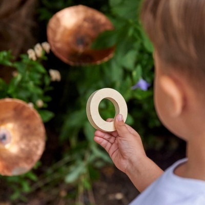 Criança segurando peça de madeira circular diante de plantas e objetos metálicos redondos