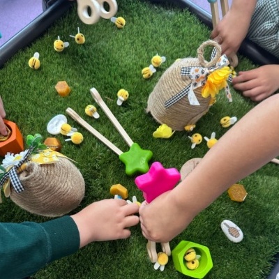 Brinquedo infantil com abelhas, potes de corda decorados e utensílios de madeira sobre relva artificial.