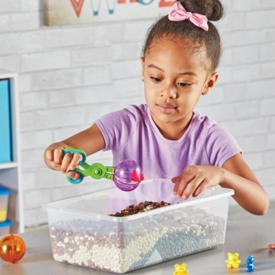 Criança com brinquedos e caixa plástica cheia de bolinhas coloridas numa mesa