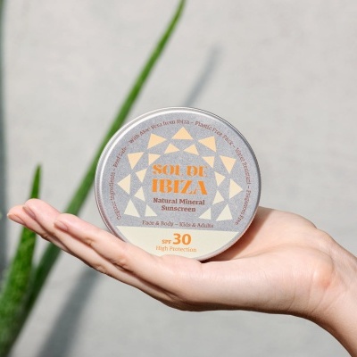 Frasco redondo de protetor solar mineral da marca SEU DA IBIZA, cor bege, na mão de uma pessoa com folhas verdes ao fundo
