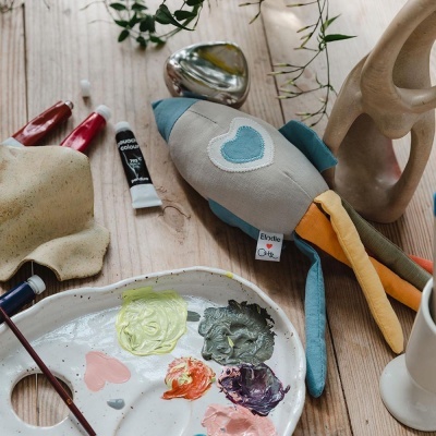 Brinquedo de pelúcia em forma de foguete, tinta e pincéis sobre mesa de madeira clara