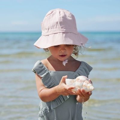 Criança na praia com chapéu rosa e vestido cinza segurando concha