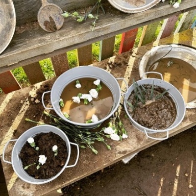 Três baldes metálicos com terra, água e barro com flores brancas sobre mesa de madeira.