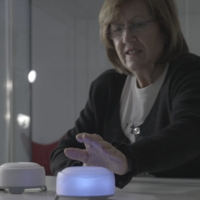 Dois dispositivos redondos em mesa branca, um iluminado em azul, com mulher interagindo.