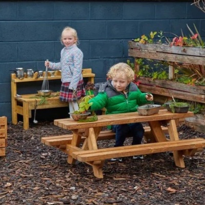 Crianças a brincar com mobiliário infantil em madeira num espaço exterior com plantas e utensílios