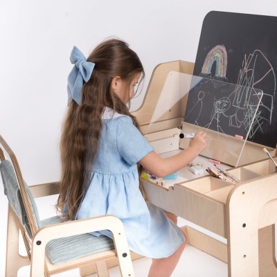 Menina a desenhar num quadro negro numa secretária de madeira clara com separador de acrílico