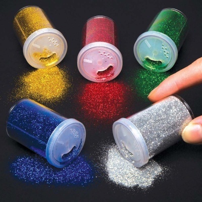 Frascos de glitter em pó nas cores amarelo, vermelho, verde, azul e prata com tampas brancas e dedo próximo ao frasco prateado.