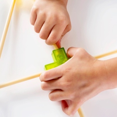  mãos infantis segurando peça plástica verde translúcida conectando varetas amarelas