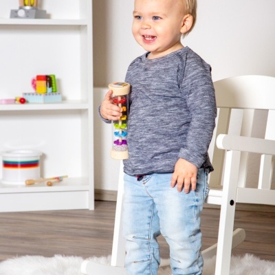 Criança em pé com camisola cinza e calças azuis segurando brinquedo colorido em ambiente interior