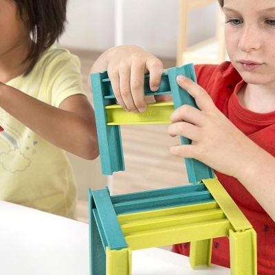 Crianças a montar cadeira colorida com peças azuis, vermelhas e amarelas numa mesa branca