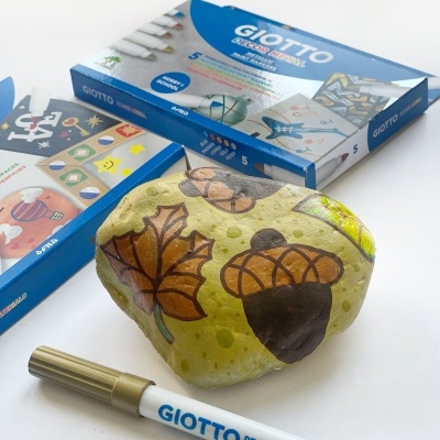 Rochas pintadas com motivos de folhas e bolotas, caneta metálica dourada Giotto e caixas de marcadores Giotto no fundo
