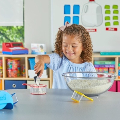 Menina com vestido azul a mexer ingredientes numa mesa com tigela transparente e copo medidor numa sala de aula colorida.