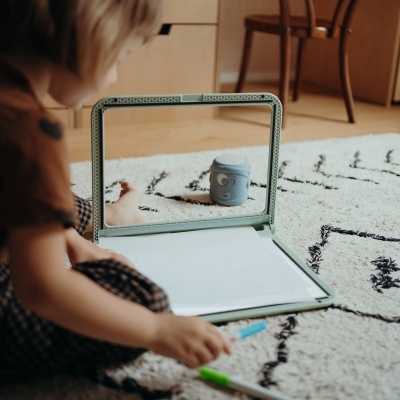 Espelho com suporte verde decorado sobre carpete branca com padrões pretos, criança a desenhar no chão.